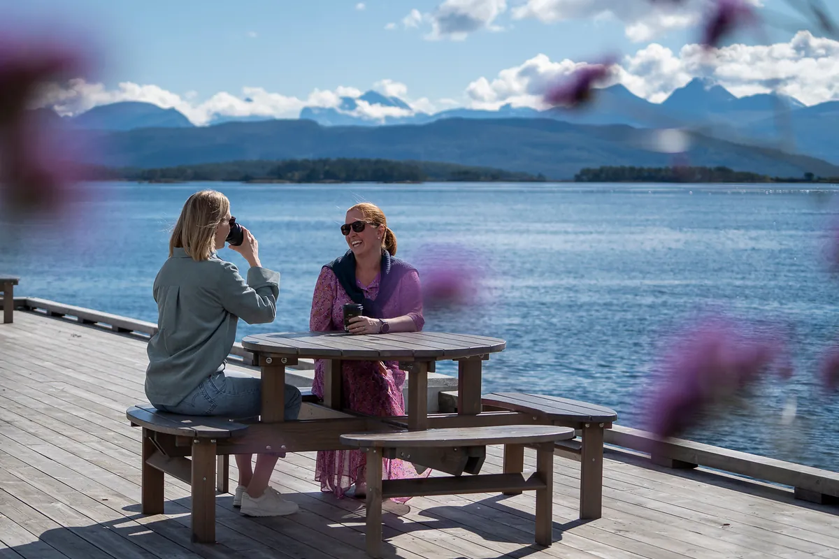 To kvinner med kaffe ved sjøen med fjellpanorama